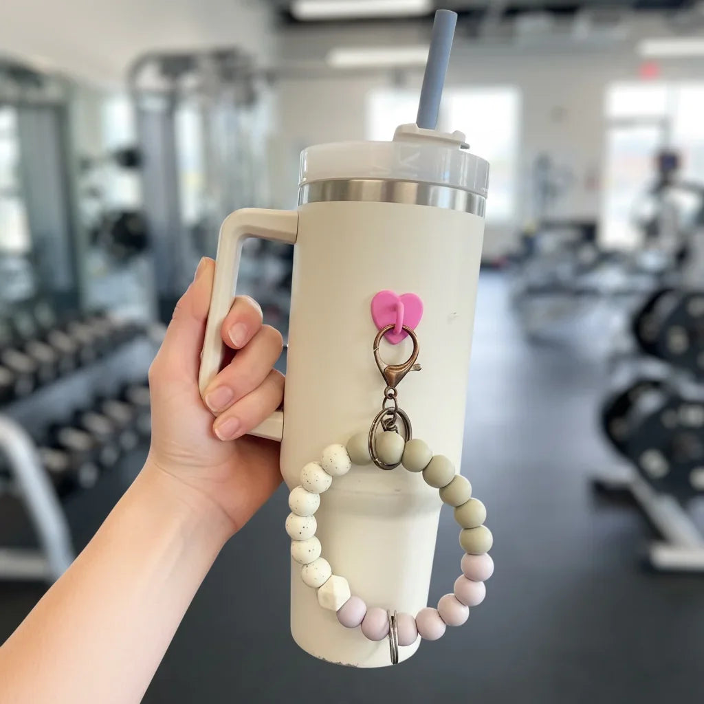 Cream travel mug with beaded handle grip and pink heart charm, held in gym setting